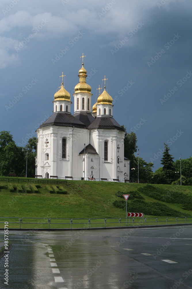 A white stone church stands on a rise near the road. White church with golden domes and crosses. A white stone church stands on a rise near the road. White church with golden domes and crosses.