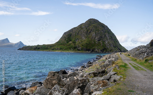 A beautiful coastal landscape showcasing a rocky shoreline with a prominent green hill rising by the sea. The clear blue water gently laps against the rocks, and the background features distant mount photo