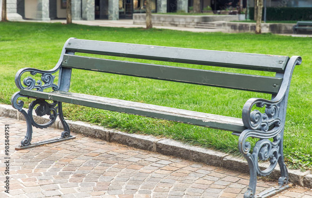 Empty park bench on cobblestone path in green park, facing green grass, trees in background, urban outdoor relaxation, public seating concept Empty park bench on cobblestone path in green park, facing green grass, trees in background, urban outdoor relaxation, public seating concept