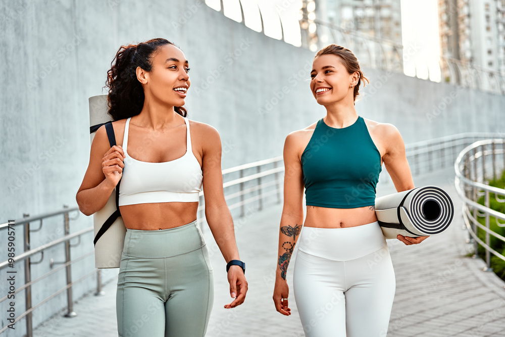 Fototapeta Attractive modern sports women walk holding mats for fitness training, yoga and joyfully chatting. Sincere friendship and sports lifestyle.