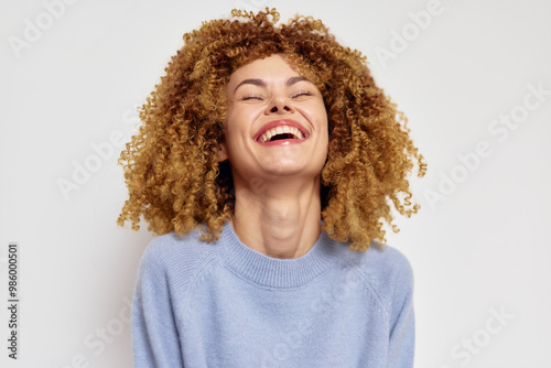 Smiling woman with curly hair, wearing a light blue sweater against a plain beige background, conveying happiness and joy with a bright expression