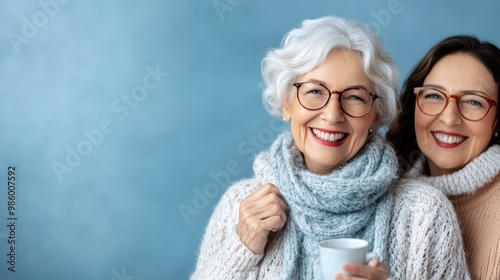 A happy elderly woman with gray hair and her daughter stand closely together, both smiling joyously while holding a white coffee mug in a cozy atmosphere.