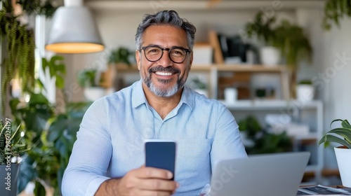 A businessman, dressed in a blue shirt, sits at a modern work desk holding a smartphone, surrounded by plants and office supplies, representing modern business communication. photo