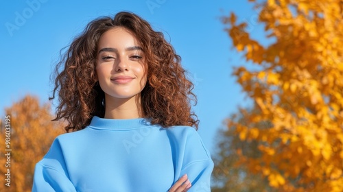 A smiling woman with flowing hair stands confidently in a blue sweater amidst vibrant autumn leaves, showcasing her beauty and the joy of the fall season. photo