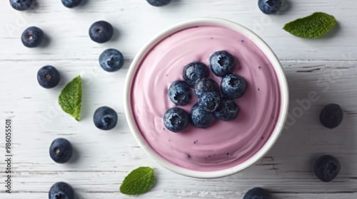 A white bowl of blueberry yogurt, placed on a clean white wooden table, with fresh blueberries sprinkled around