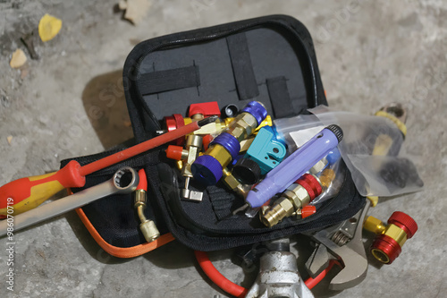Black tool bag filled with various HVAC tools and components, including gauges, hoses, a screwdriver, and other specialized fittings. The bag is placed on a concrete surface. Used for HVAC repair work