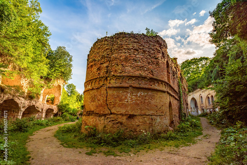 Crumbling brick walls of Tarakaniv Fort, an abandoned 19th-century fortress in Ukraine. Overgrown with vegetation, the circular tower stands amid dirt paths and arched structures. photo