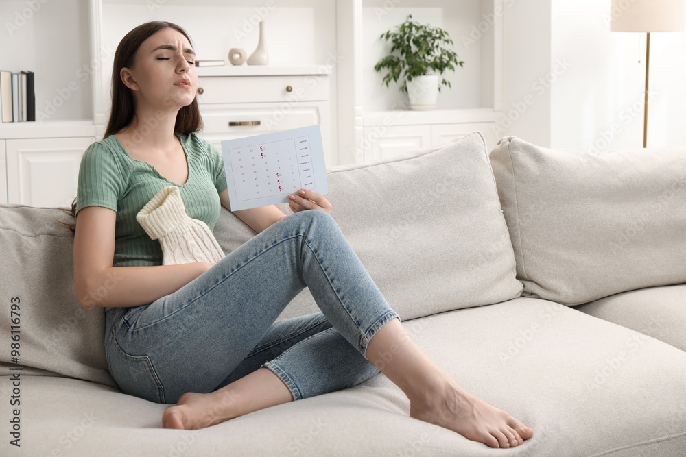 Woman holding calendar with marked menstrual cycle days and using hot water bottle on sofa at home Woman holding calendar with marked menstrual cycle days and using hot water bottle on sofa at home