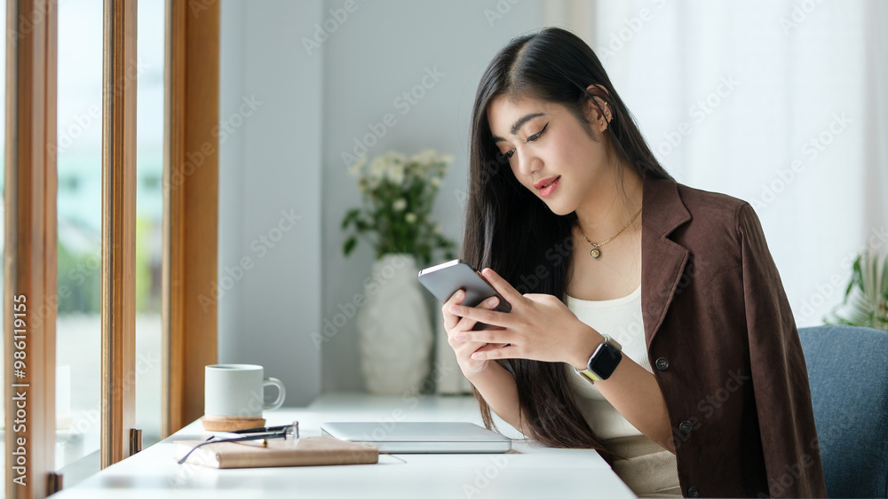 Woman with long dark hair is sitting at a bright, modern desk by a large window, holding a smartphone and smiling as she engages with her device. Woman with long dark hair is sitting at a bright, modern desk by a large window, holding a smartphone and smiling as she engages with her device.