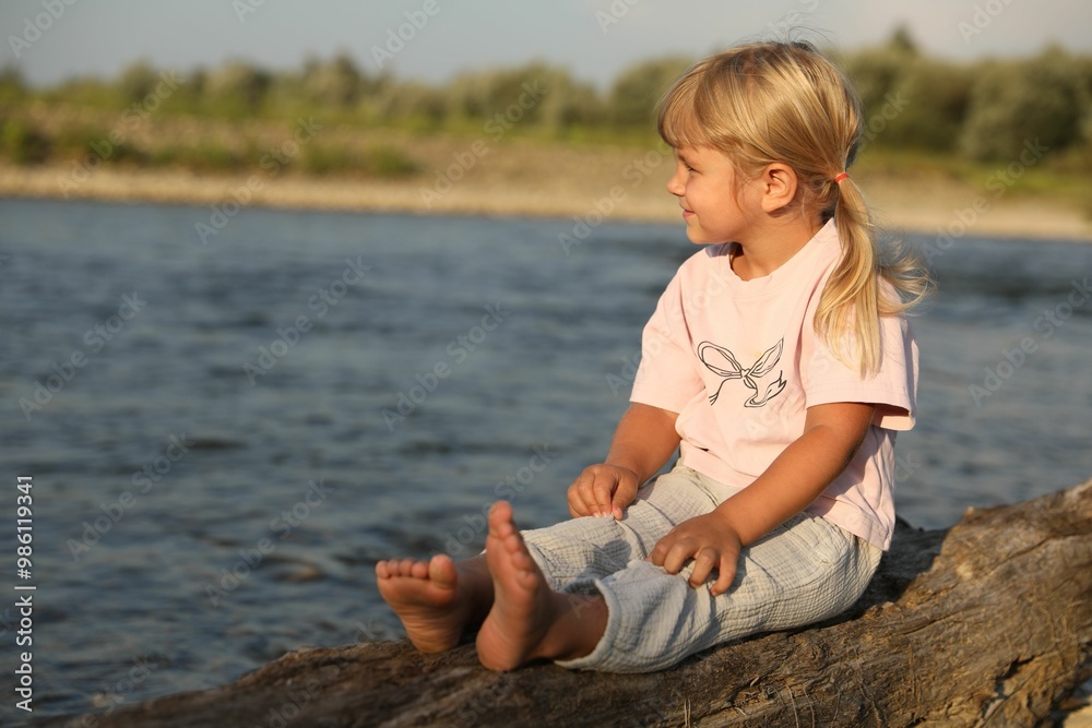 Cute little girl sitting on tree trunk near river, space for text. Child enjoying beautiful nature Cute little girl sitting on tree trunk near river, space for text. Child enjoying beautiful nature
