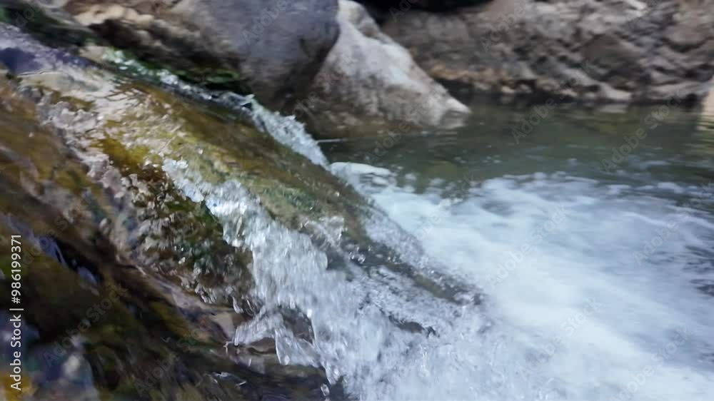A slow-motion video capturing a small waterfall in a mountain stream. The water cascades gently over rocks, with each droplet and ripple accentuated, creating a tranquil and calming scene in nature. A slow-motion video capturing a small waterfall in a mountain stream. The water cascades gently over rocks, with each droplet and ripple accentuated, creating a tranquil and calming scene in nature.