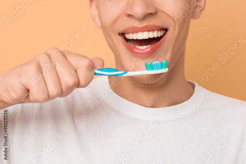 Young man brushing his teeth on beige background, closeup