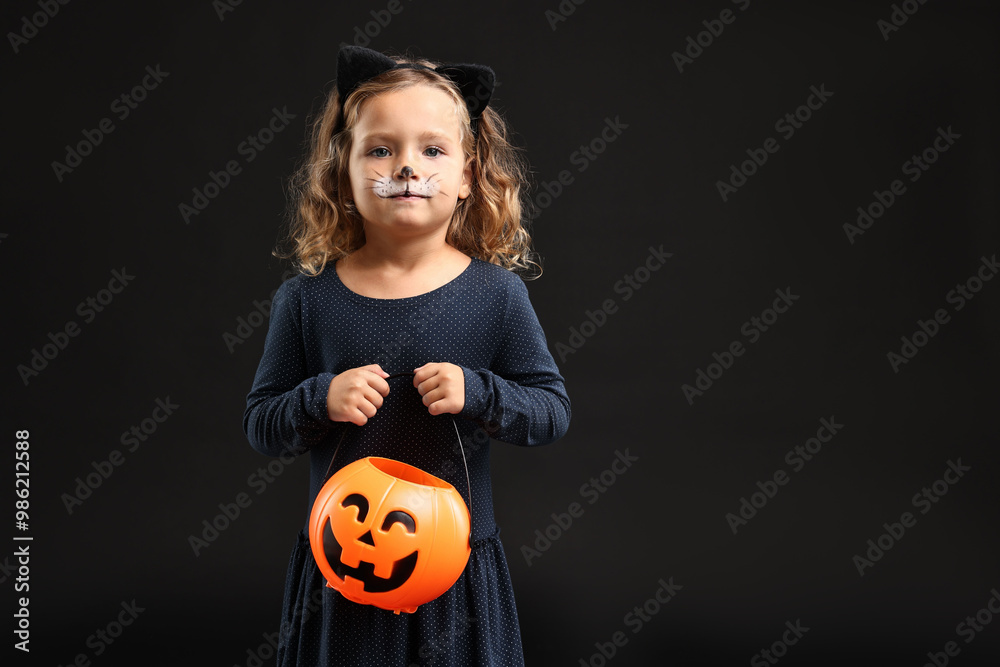 Fototapeta premium Cute girl with pumpkin bucket dressed like cat for Halloween celebration on black background, space for text