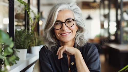 A smiling woman with gray hair and glasses poses in a stylish, modern environment, exuding confidence and timeless beauty.