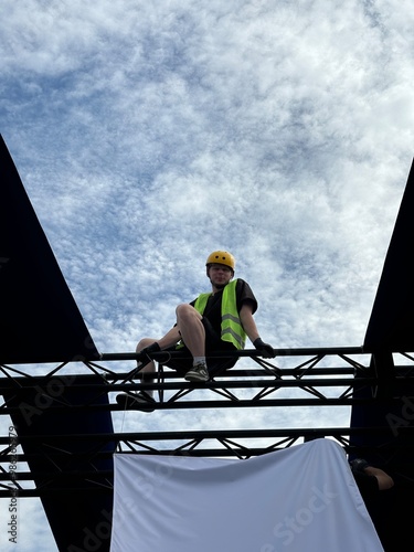 High-altitude worker wearing a green safety vest and yellow helmet, seated on a metal framework. This image emphasizes construction safety and professionalism in the field. Vertical photo photo