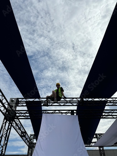 Young construction worker in a green vest and yellow helmet sitting on a high metal structure, showcasing safety and expertise in the industry. Professional work attire and gloves. Vertical photo photo