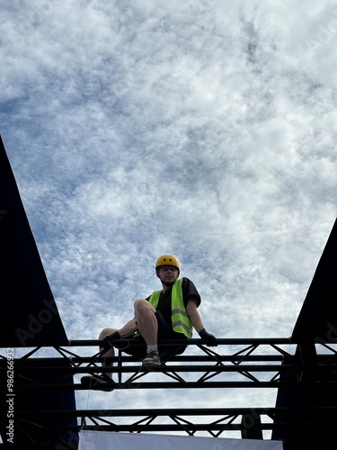Obraz na płótnie Focused young male construction worker in a green vest and yellow helmet, sitting confidently on a tall metal structure. A representation of safety and skill in high-rise construction. Vertical photo w salonie