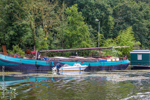 Old barge used as houseboat on bank of shipping canal outside Maastricht city, small boat, water with aquatic plants on surface, leafy trees on background, cloudy day in South Limburg, Netherlands