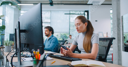 Wallpaper Mural Beautiful girl spending time on phone while sitting at workplace in office. Corresponding with clients or checking her mail. Using computer and smartphone for work efficiency. Modern office. Torontodigital.ca