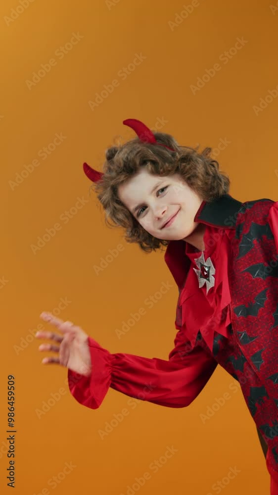 Medium vertical shot of mischievous Caucasian boy with blond curly hair, in devil outfit, with horns appearing from corner on orange background, waving then disappearing. Copy space, template Medium vertical shot of mischievous Caucasian boy with blond curly hair, in devil outfit, with horns appearing from corner on orange background, waving then disappearing. Copy space, template