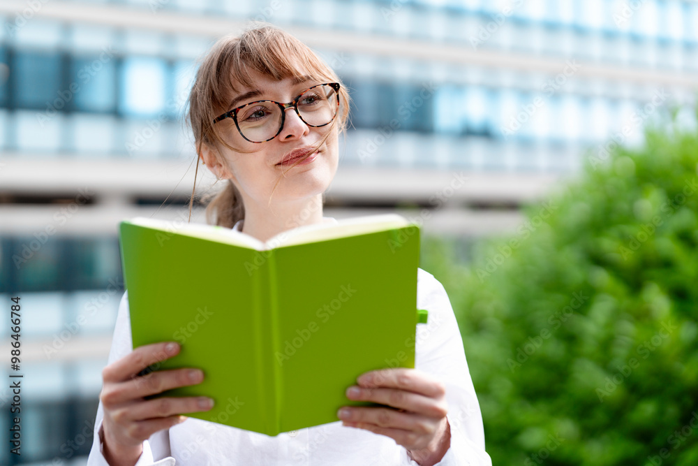Joyful Woman Reading Green Book Outdoors in Modern Cityscape During a Sunny Day Joyful Woman Reading Green Book Outdoors in Modern Cityscape During a Sunny Day