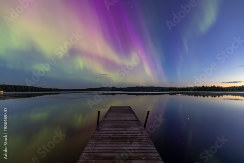 A beautiful Northern Lights is shown in the night sky, with vibrant colors such as pink, green, and purple.  In the foreground, there is a wooden pier jutting out into the water.