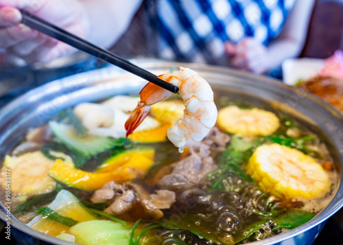 Chopsticks grabbing shrimp in a boiling shabu with a woman's hand in the blurred background