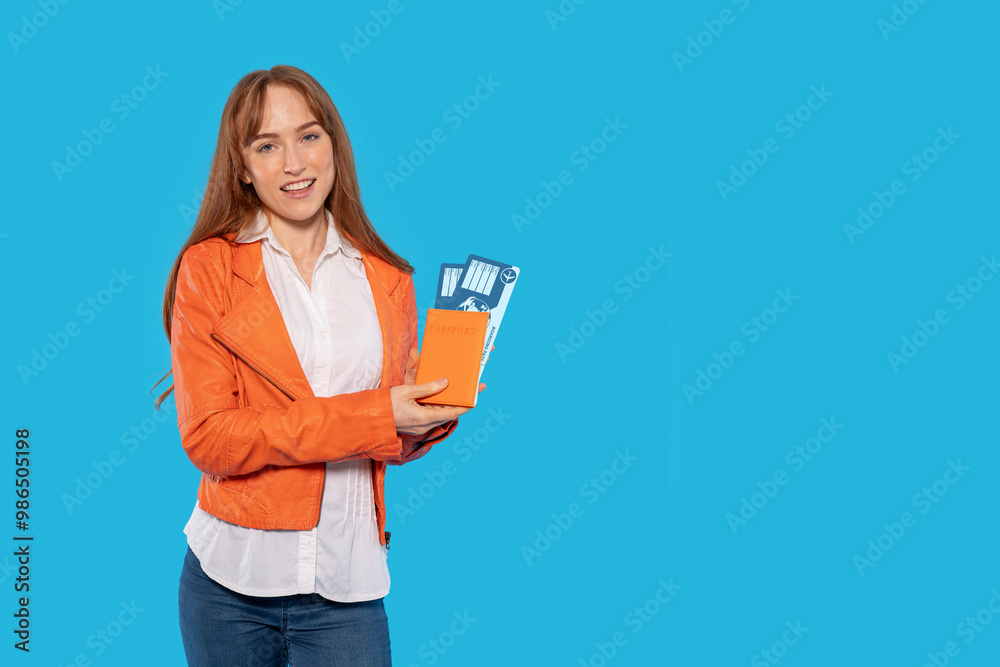 Young Woman in Orange Jacket Smiles With Boarding Pass and Travel Guide Against a Blue Backdrop Young Woman in Orange Jacket Smiles With Boarding Pass and Travel Guide Against a Blue Backdrop