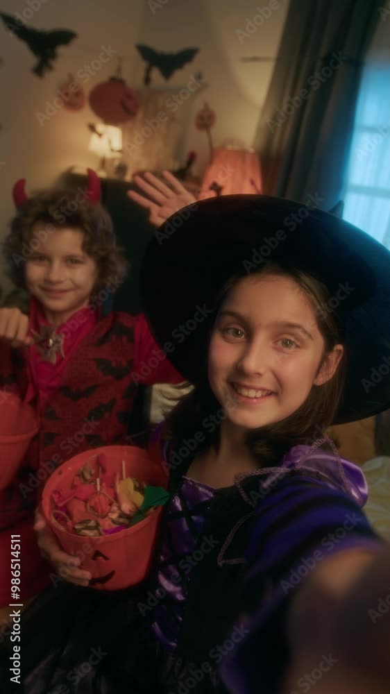 Medium vertical UGC shot of girl in witch dress and boy in devil outfit sitting together with buckets of Halloween candy, smiling, waving while taking selfie or filming video on smartphone Medium vertical UGC shot of girl in witch dress and boy in devil outfit sitting together with buckets of Halloween candy, smiling, waving while taking selfie or filming video on smartphone