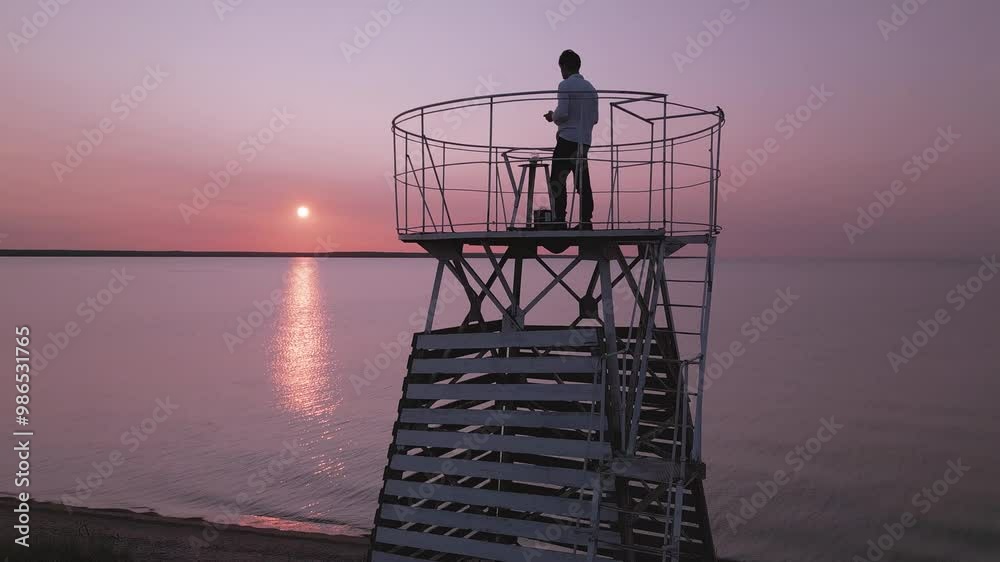 Male model with white shirt standing on white lighthouse near sea at red-pink sunset Male model with white shirt standing on white lighthouse near sea at red-pink sunset