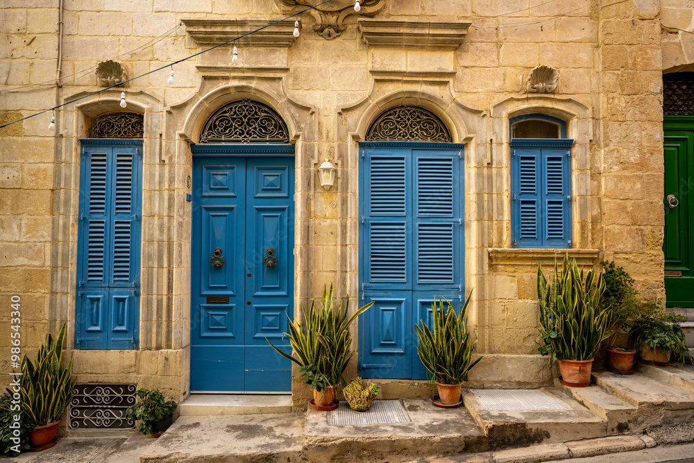 Fototapeta premium Charming facade with vibrant blue wooden doors and shutters, set against traditional stone walls in Malta. Potted plants add a touch of greenery to this picturesque Mediterranean scene