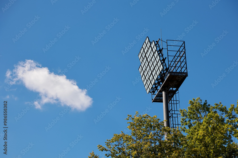 A Tall Floodlight Tower Against A Clear Blue Sky With A Single White Cloud, Surrounded By Tree Foliage. Represents Sports And Outdoor Events. A Tall Floodlight Tower Against A Clear Blue Sky With A Single White Cloud, Surrounded By Tree Foliage. Represents Sports And Outdoor Events.