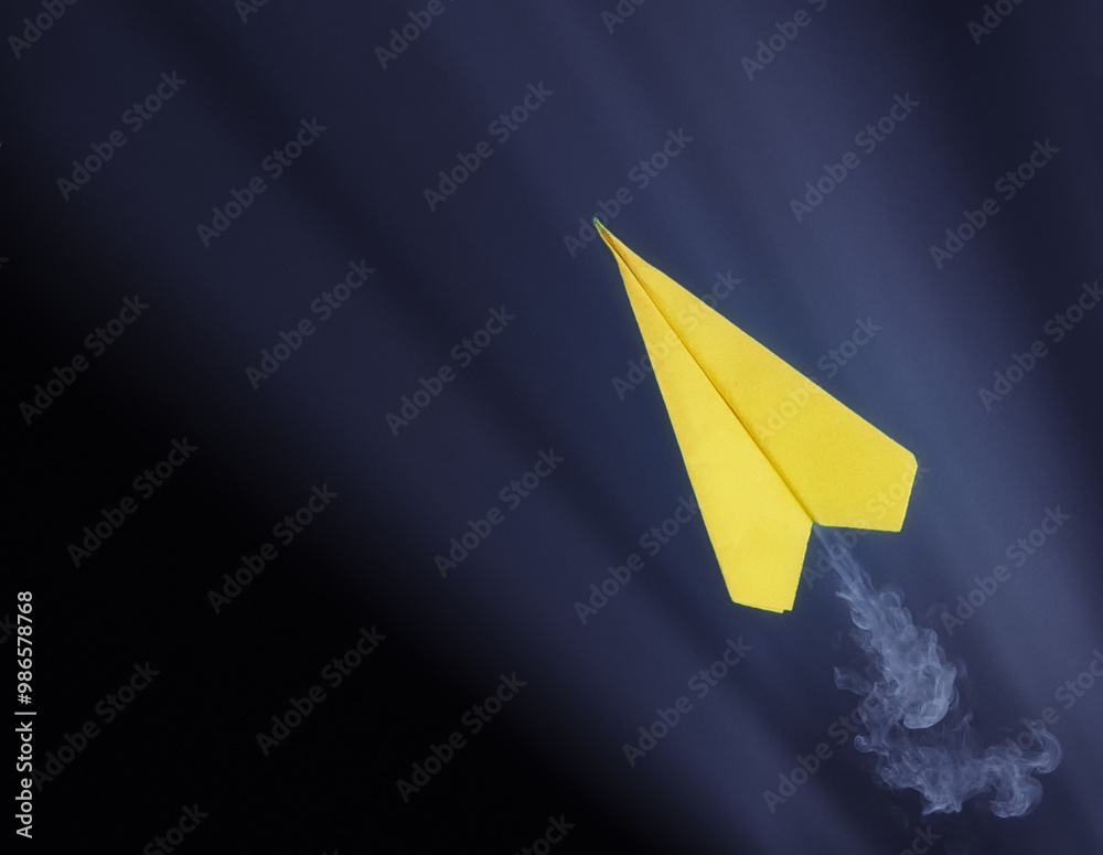 Yellow paper airplane with smoke trail flying against dark background.