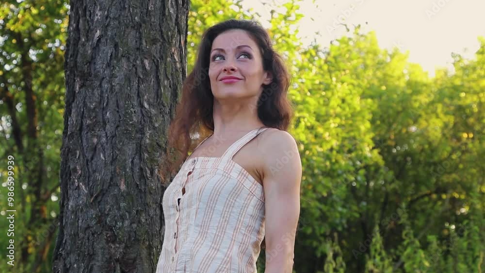 Beautiful long hair happy smiling woman enjoying the sunny summer day near the tree, carefree touching the big tree trunk the hands Beautiful long hair happy smiling woman enjoying the sunny summer day near the tree, carefree touching the big tree trunk the hands