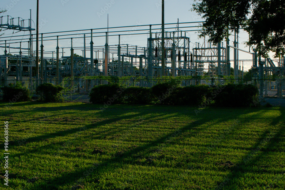 Electric Substation Framed with tree limbs Low Wide view across bright green grass and trees late afternoon sun with long shade. Near sunset with green bushes in front of the fence. No people and room Electric Substation Framed with tree limbs Low Wide view across bright green grass and trees late afternoon sun with long shade. Near sunset with green bushes in front of the fence. No people and room