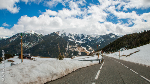 A winding mountain road leads through a snowy landscape towards a ski resort nestled in the Pyrenees mountains under a bright blue sky.