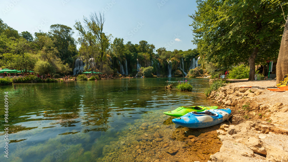 Αυτοκόλλητο Kayaks by tranquil riverbank with scenic waterfall backdrop, perfect for summer adventure