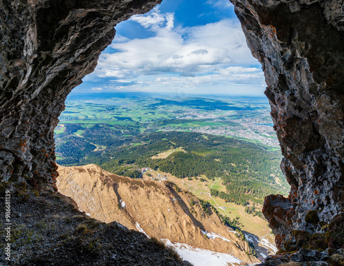 A mountain cave frames a breathtaking panorama of rolling hills, verdant valleys, and distant towns, offering a unique perspective on the Swiss Alps' grandeur and beauty.