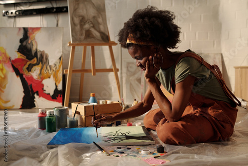 Fototapeta Naklejka Na Ścianę i Meble -  Artist sitting on floor painting with various art supplies around. Abstract and portrait paintings on easel and wall in background enhancing creative atmosphere