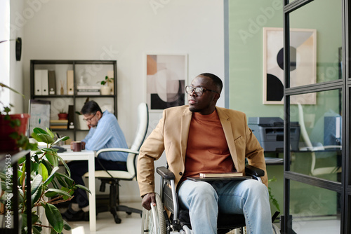 Person in wheelchair working at desk in light-filled office next to colleague sitting at separate desk concentrating on task photo
