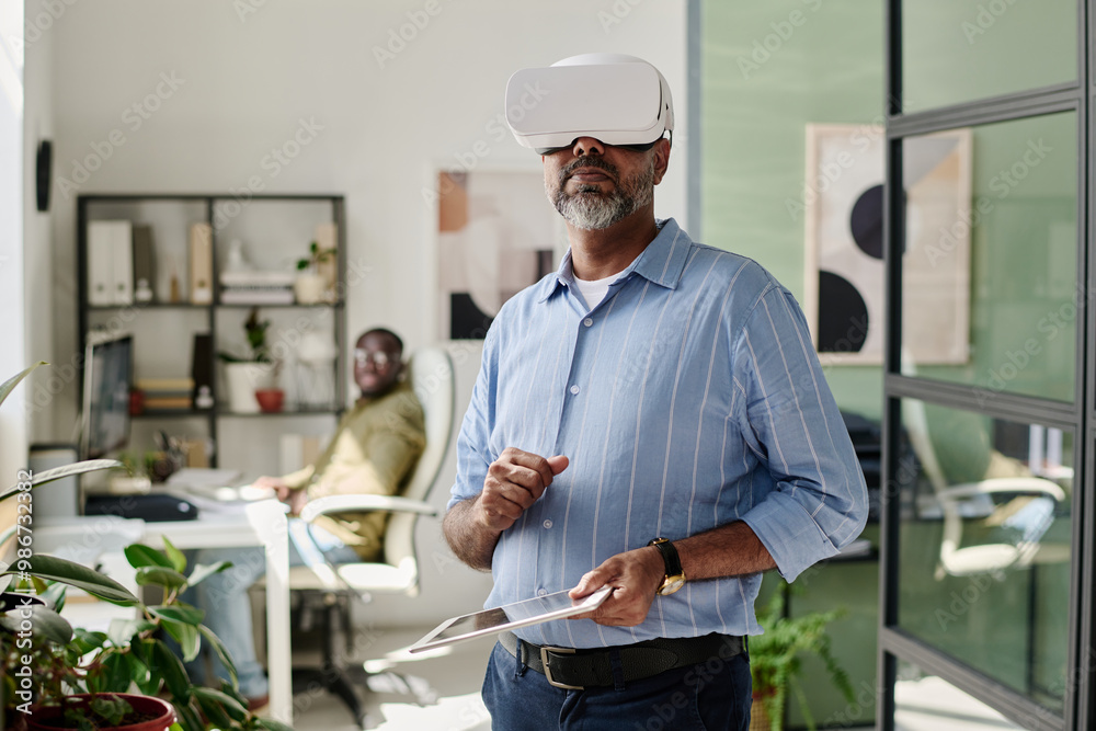 Man engaging with virtual reality headset in modern office setting, standing near desk with colleague in background, holding clipboard, and interacting with VR Man engaging with virtual reality headset in modern office setting, standing near desk with colleague in background, holding clipboard, and interacting with VR