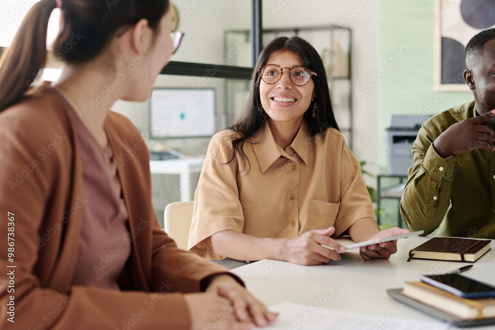 Smiling woman wearing glasses engaging colleagues at meeting table in modern office with large windows. Multiracial team collaborating in workspace filled with natural light and technology