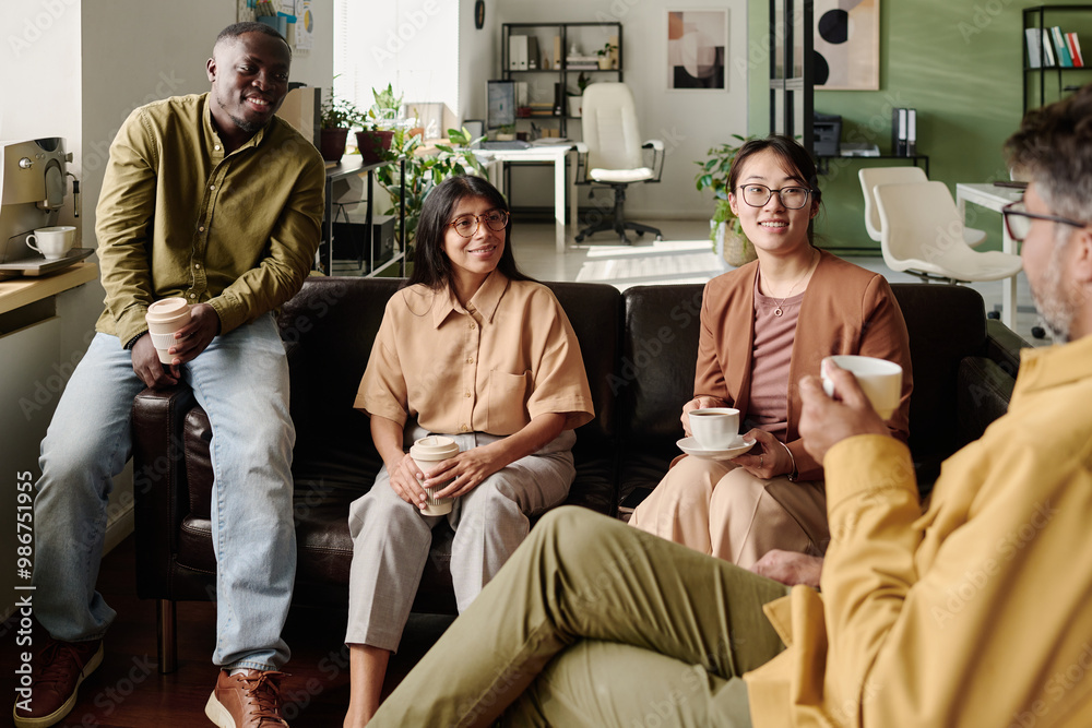 © pressmaster - Group of diverse coworkers sitting on a couch and exchanging ideas in modern office with relaxed atmosphere, holding coffee cups and engaging in conversation