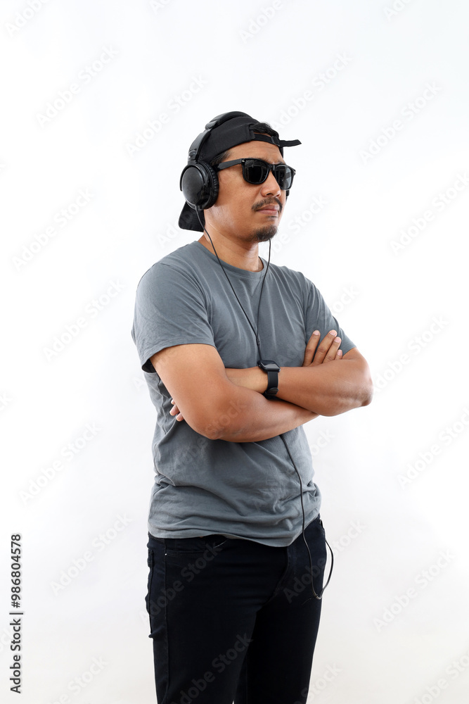 Fototapeta premium Asian man enjoying his favorite music. Happy stylish man in black hat and sunglasses with headphones listening and smiling while standing against white background