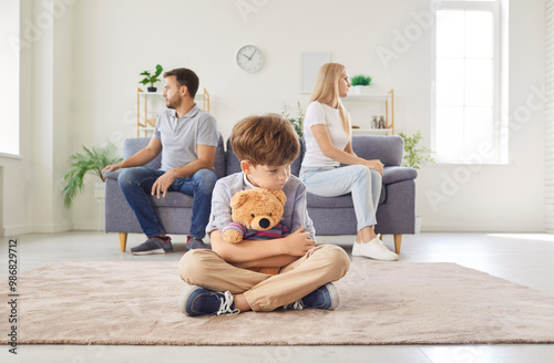 Frustrated upset boy son sitting on floor with toy bear while his mother and father sitting on opposite sides of sofa ignoring each other after quarrel at home. Divorce and family conflicts concept. photo