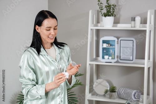 Young woman with jar of cream and cosmetic refrigerator in bathroom