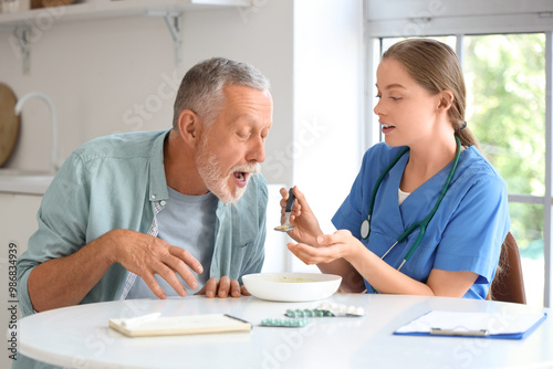 Nurse feeding senior man suffering from Parkinson syndrome at table in kitchen