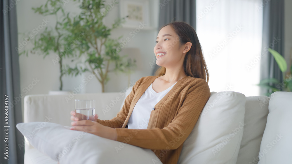 Happy young Asian woman drinking water while sitting on the sofa in the living room Happy young Asian woman drinking water while sitting on the sofa in the living room