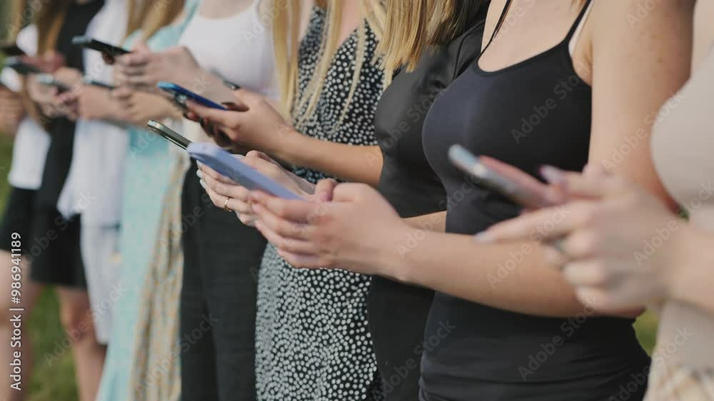 Group of young women are standing in a line outdoors, each looking down at their smartphones.