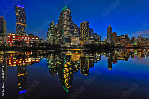 A city of Austin skyline illuminated at night is reflected perfectly in calm water of the Colorado River below. 